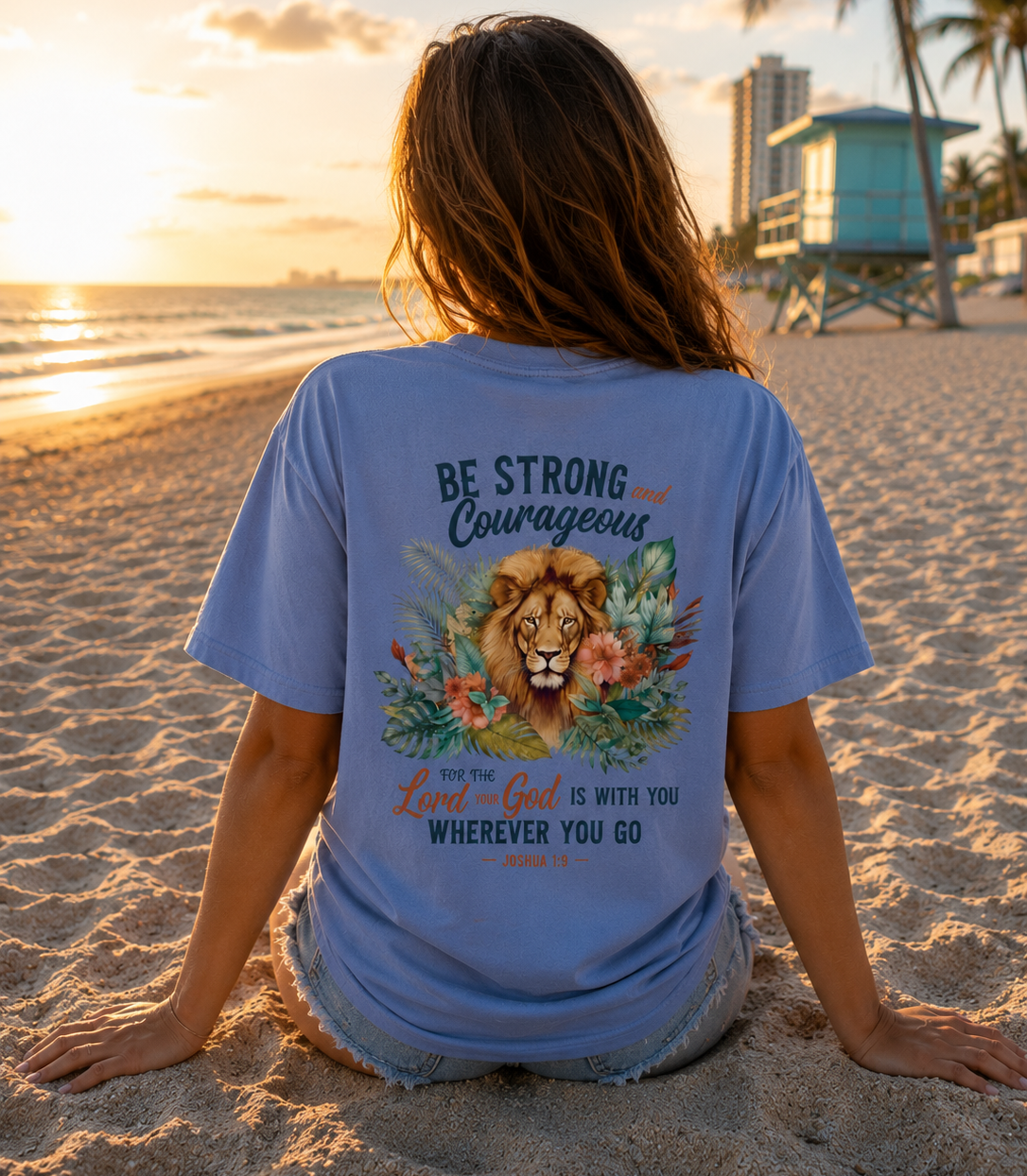 Person wearing a blue t-shirt with a lion graphic and bible verse on a beach at sunset.