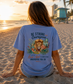 Person wearing a blue t-shirt with a lion graphic and bible verse on a beach at sunset.