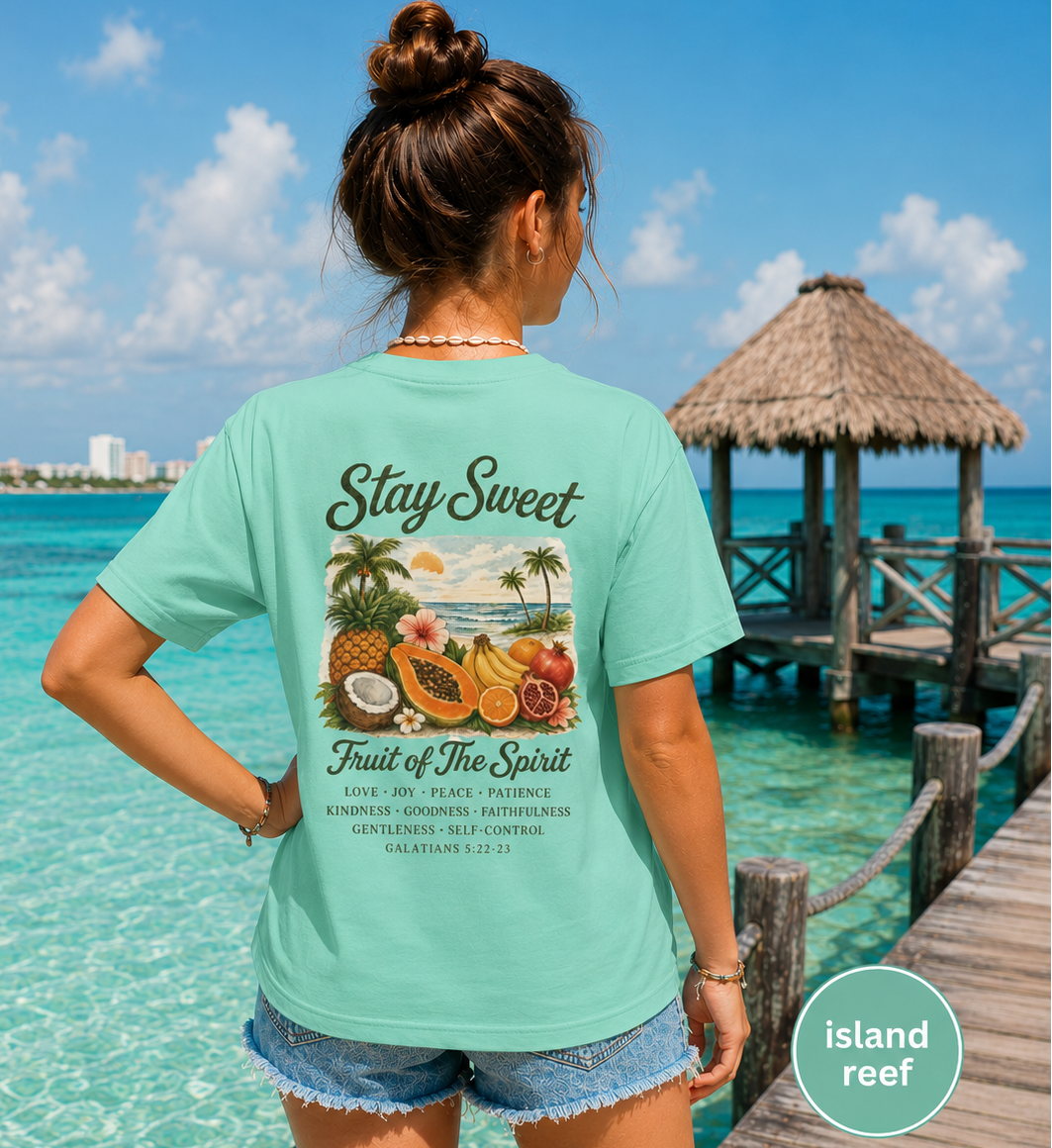 Woman wearing a teal t-shirt with a fruit design and text by a tropical beach.