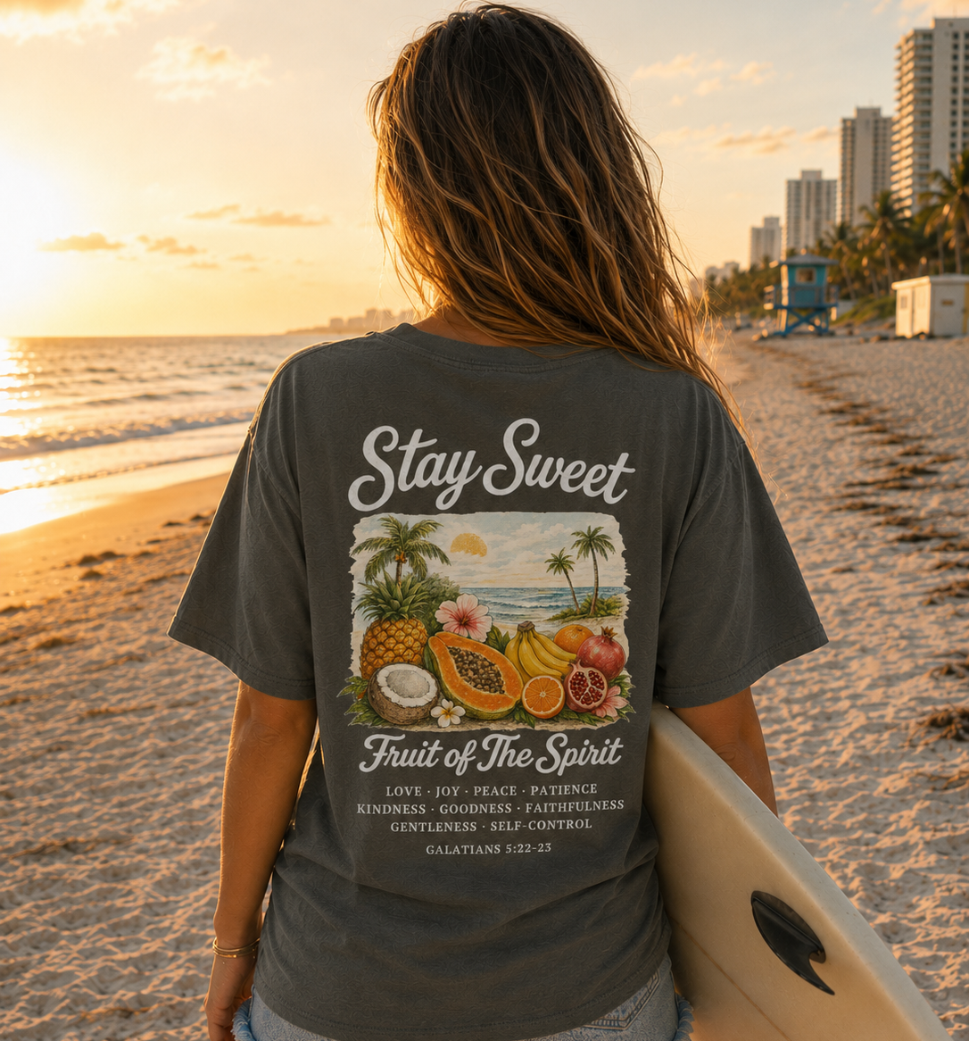 Person on a beach holding a surfboard wearing a 'Stay Sweet' t-shirt with fruit of the spirit bible verse