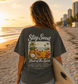 Person on a beach holding a surfboard wearing a 'Stay Sweet' t-shirt with fruit of the spirit bible verse