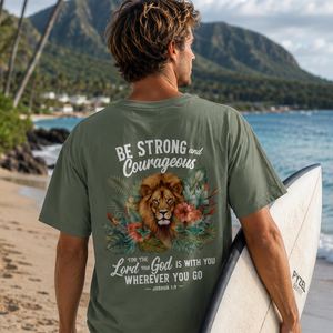 Man holding a surfboard on a beach with a scenic background, wearing a Christian t-shirt with a motivational design.