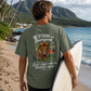 Man holding a surfboard on a beach with a scenic background, wearing a Christian t-shirt with a motivational design.