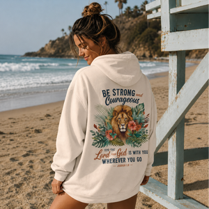 A woman stands on a beach facing away from the camera, wearing a cream-colored hoodie with a large lion and floral graphic on the back, accompanied by the text 'BE STRONG and Courageous' and 'the Lord and God IS WITH YOU WHEREVER YOU GO'.